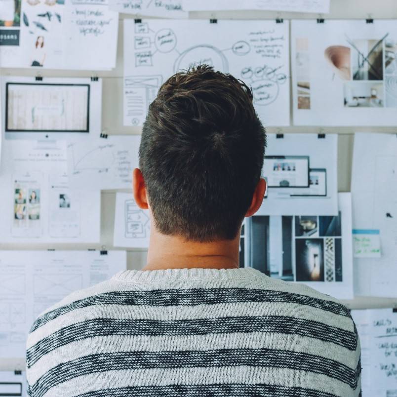 A person with short brown hair, wearing a white and gray striped sweater, is seen from behind while examining a wall covered with papers, charts, and diagrams related to PMP process groups.