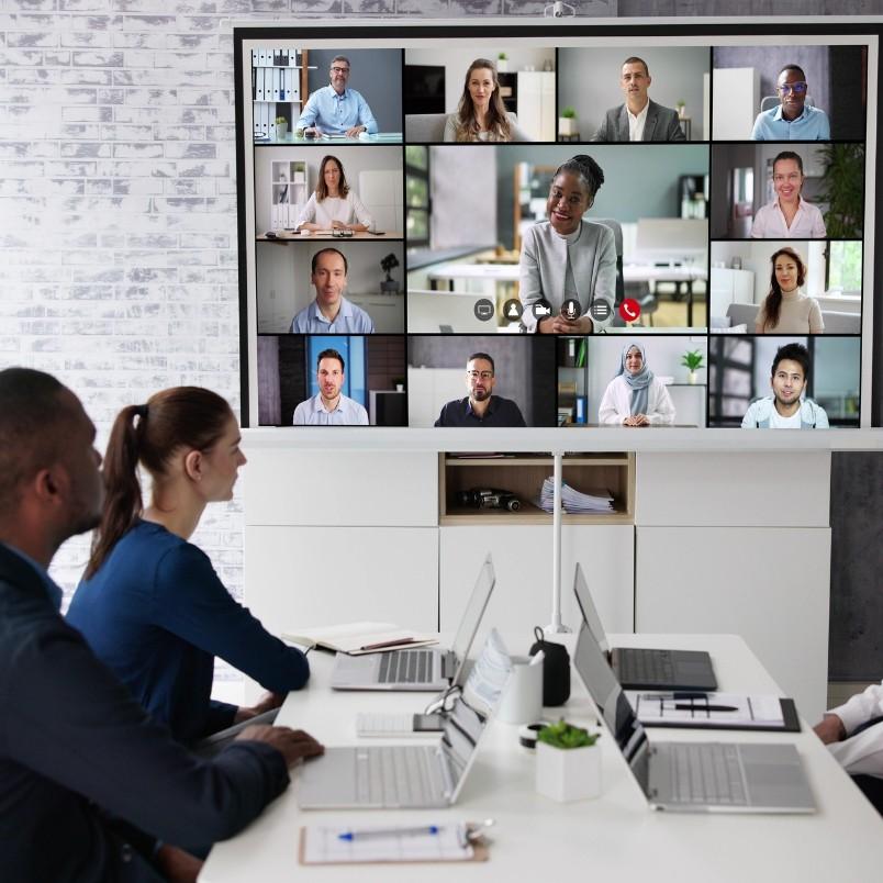 Four people sit at a conference table with laptops, showcasing agile team collaboration in a hybrid work model as they join a video call with many others whose faces are displayed on a large screen at the front of the room.