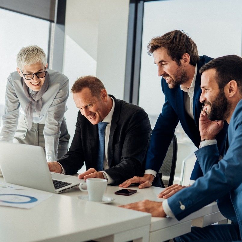 Four business professionals, three men and one woman, gathered around a laptop in a bright office, smiling as they discuss generative AI in project management. A coffee cup and papers are on the table.