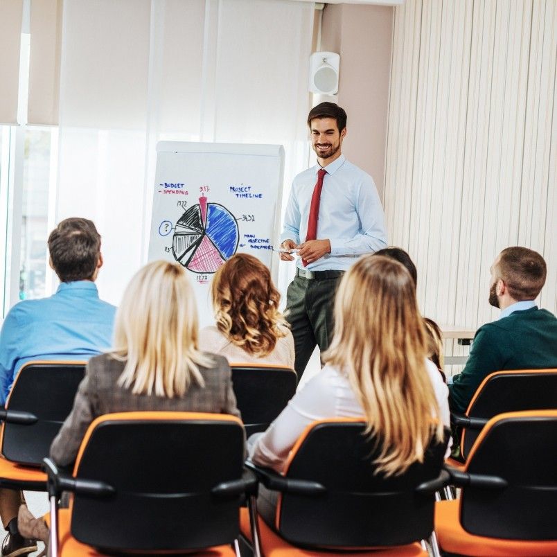 A man in business attire stands near a flip chart with pie and bar graphs, presenting project governance insights to a seated audience in an office setting. The attendees face him, listening attentively.
