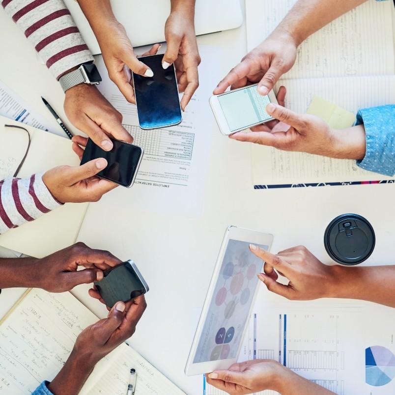 An agile team collaborates in a hybrid work model, gathered around a table with smartphones, tablets, notebooks, documents, and a coffee cup in view.