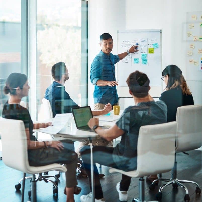 A man demonstrates leadership as he stands by a whiteboard, presenting to four colleagues seated around a table with laptops and papers in a modern office with glass walls. Sticky notes are visible on the whiteboard and wall.