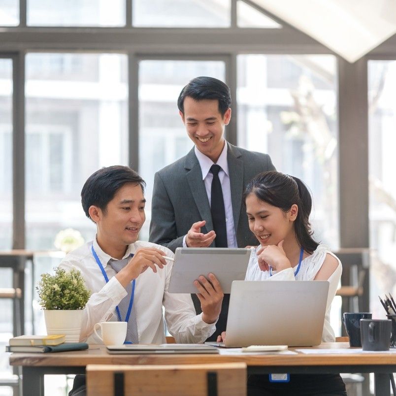 Three business professionals in an office collaborate around a tablet, discussing generative AI in project management. Two are seated, one stands behind, all smiling and engaged. Laptops and documents are on the table, with bright windows in the background.