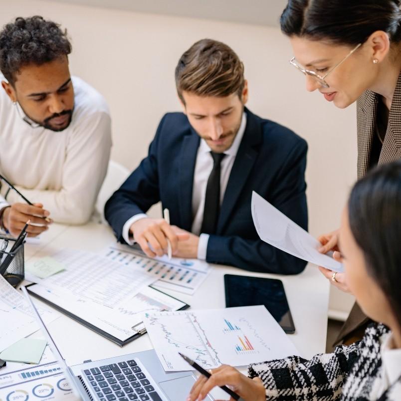 Four professionals collaborate at a desk with charts, graphs, and a laptop, showcasing agile team collaboration in a hybrid work model as they discuss documents and financial data in a bright office environment.