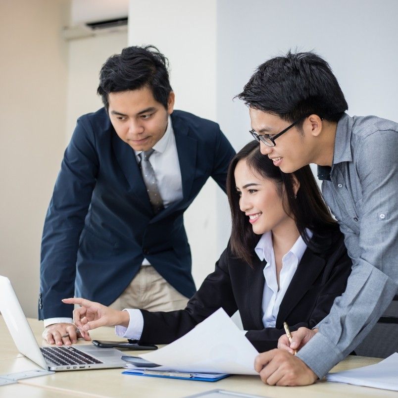 Three young professionals, two men and one woman, are gathered around a laptop. The woman is seated, smiling and pointing at the screen as they discuss generative AI in project management; the men stand beside her, all appearing engaged.