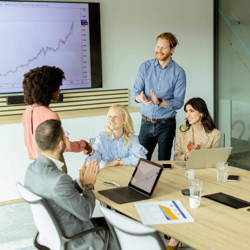 A group of five colleagues in a modern office applaud as two people shake hands. Laptops, papers, and charts related to generative AI in project management are on the table, while a large display shows a rising graph, reflecting a positive, collaborative mood.