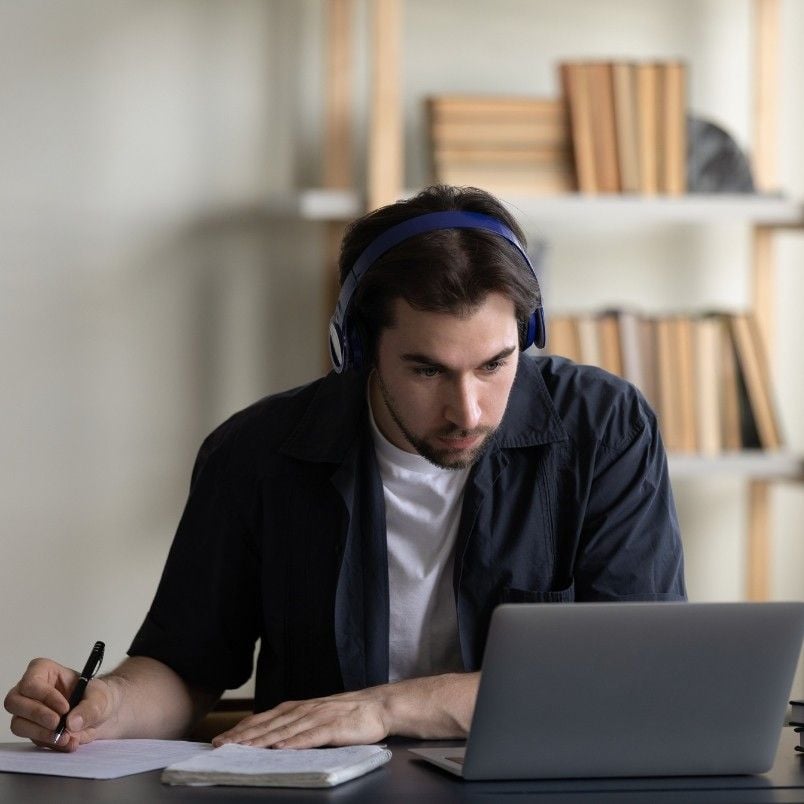 A young man wearing headphones sits at a desk, focused on researching PMP certification cost on his laptop while taking notes in a notebook. Bookshelves filled with books are visible in the background.