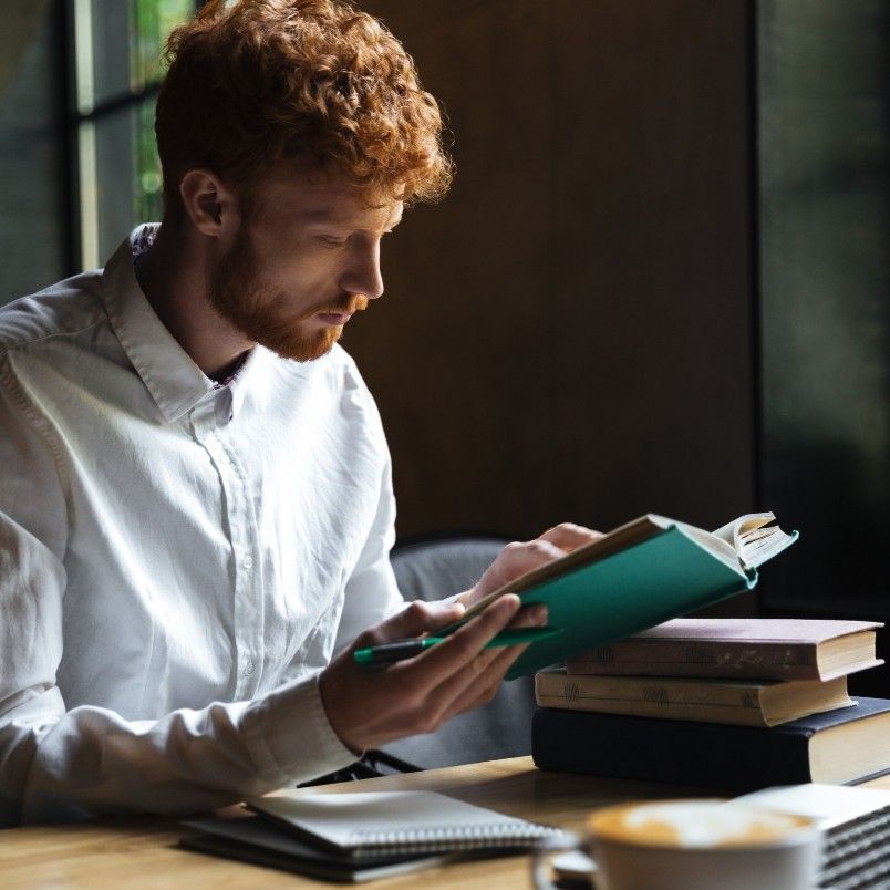 A young man with curly red hair and a white shirt sits at a table reading a book on how to get through CAPM. He holds a pen, with an open notebook in front of him, a stack of books nearby, and a cup in the foreground.