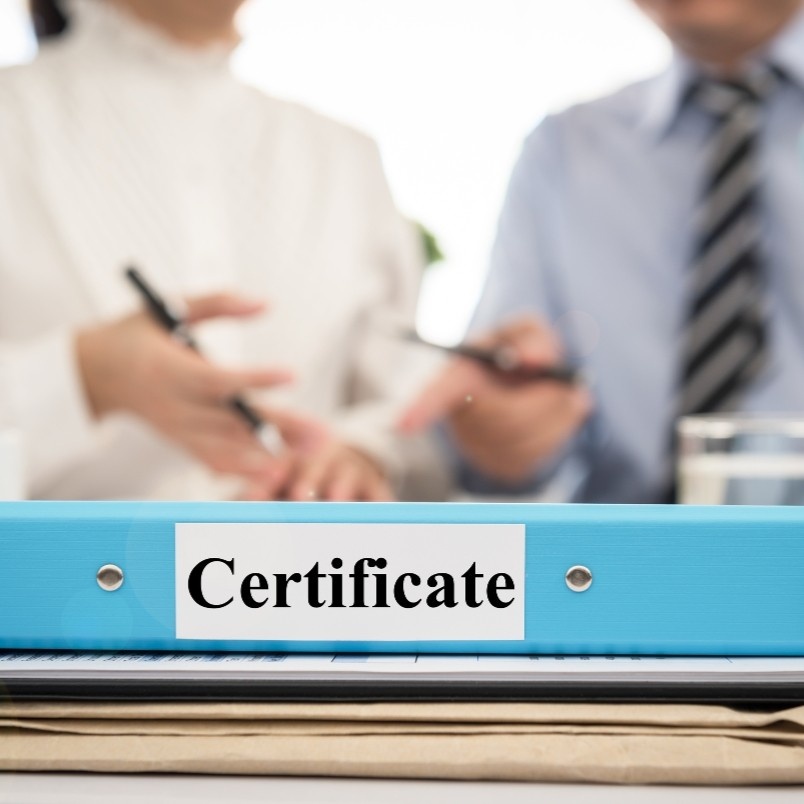 A blue binder labeled "Certificate" sits on a desk, hinting at CAPM certification requirements, while two people in business attire are blurred in the background, appearing to discuss documents.