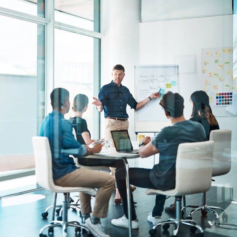 A man stands by a whiteboard giving a CAPM course presentation to four seated colleagues in a modern conference room with large windows. Laptops and documents are on the table.