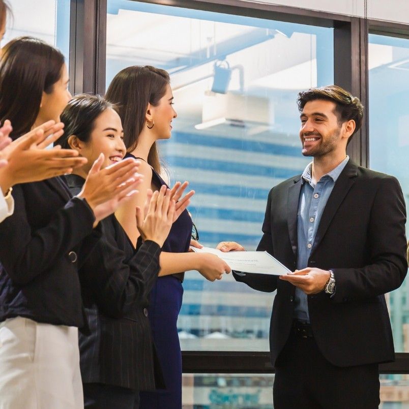 A smiling man in a suit hands a certificate to a woman in a blue dress, celebrating her achievement of meeting CAPM certification requirements, while others applaud in an office with large windows in the background.