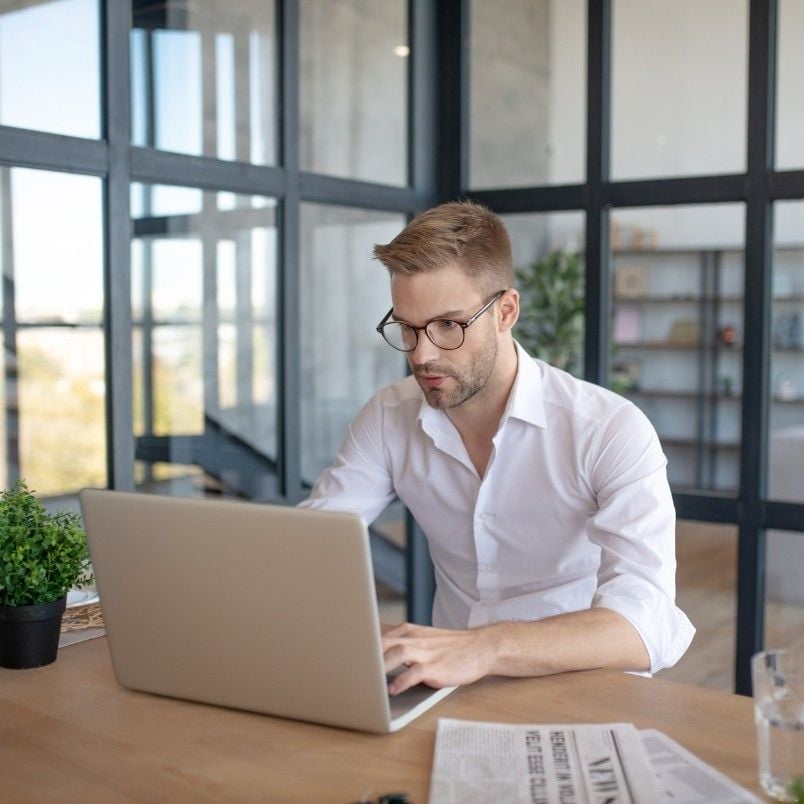 A certified associate in project management, wearing glasses and a white shirt, works on a laptop at a desk in a modern office. A newspaper, notebook, pen, and potted plant are on the desk. Large glass windows and shelves are in the background.