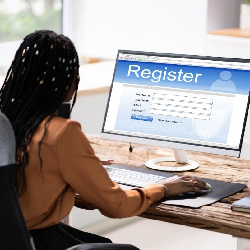 A person with long braided hair sits at a desk using a computer, filling out an online registration form on how to get through CAPM, entering first name, last name, email, and password.