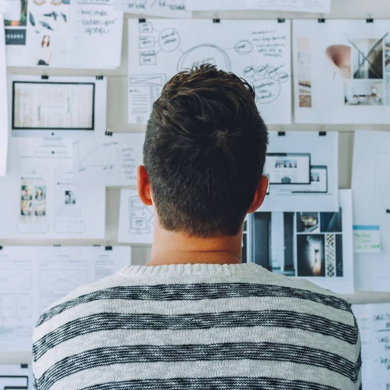 A person with short brown hair, wearing a white and gray striped sweater, is looking at a wall covered in papers, sketches, and pmp exam prep materials, suggesting brainstorming or planning.