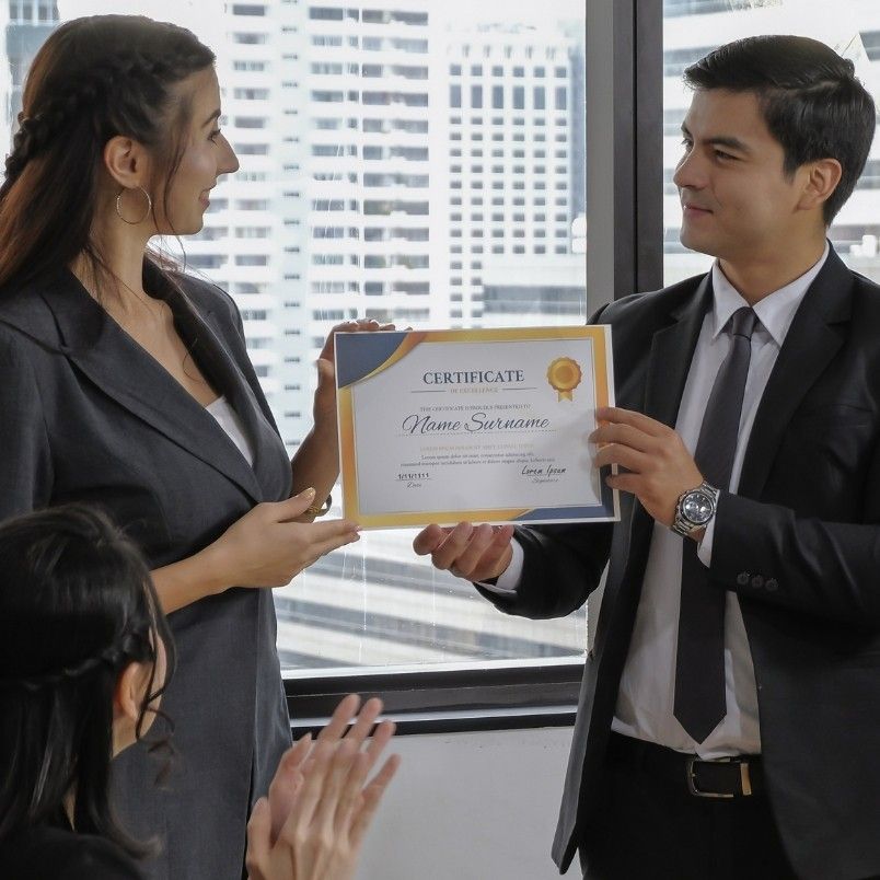 A man in a suit hands a CAPM cert to a woman in business attire while two colleagues seated nearby applaud. They are in an office with large windows and a cityscape view.