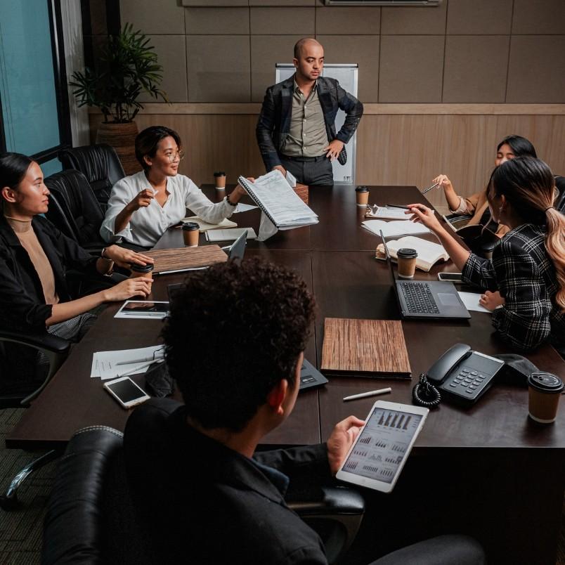 A group of six people in business attire meet around a conference table with laptops, papers, coffee cups, and charts, as one person stands and addresses the group about project governance.