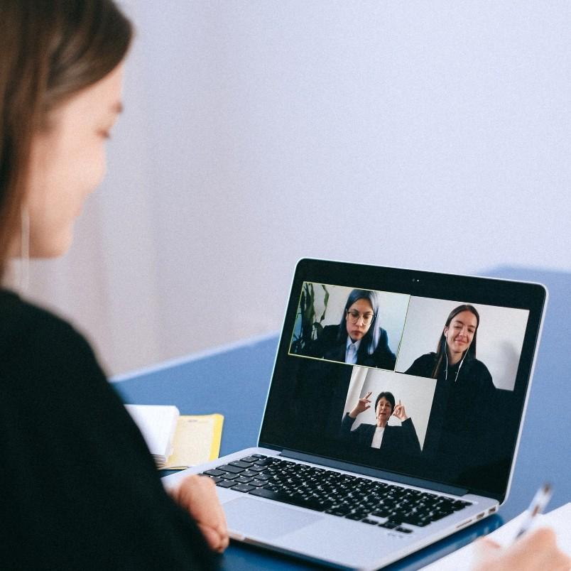 A woman sits at a desk, participating in a video call on her laptop with three colleagues. She has a notebook and pen nearby, highlighting agile team collaboration in a hybrid work model.