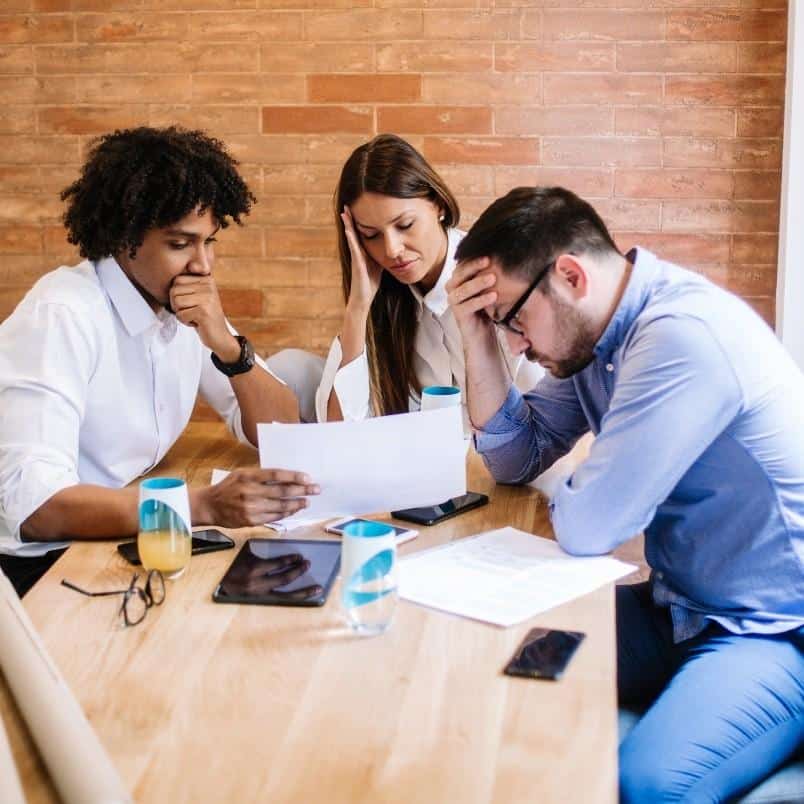 Three people sit at a table, looking stressed while reviewing documents. With drinks, phones, and tablets before them, they show how emotional intelligence in project management is vital when handling challenging situations.