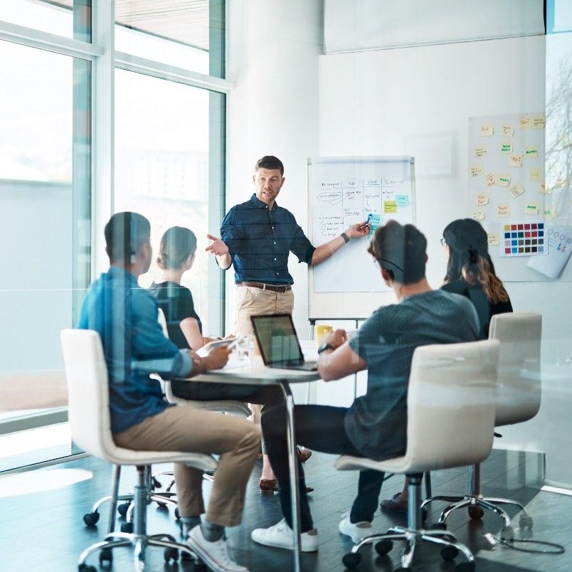 A man stands and presents on project governance to four seated colleagues in a glass-walled office, using a flip chart and notes on the wall. Laptops and notebooks are on the table. Bright daylight fills the modern workspace.