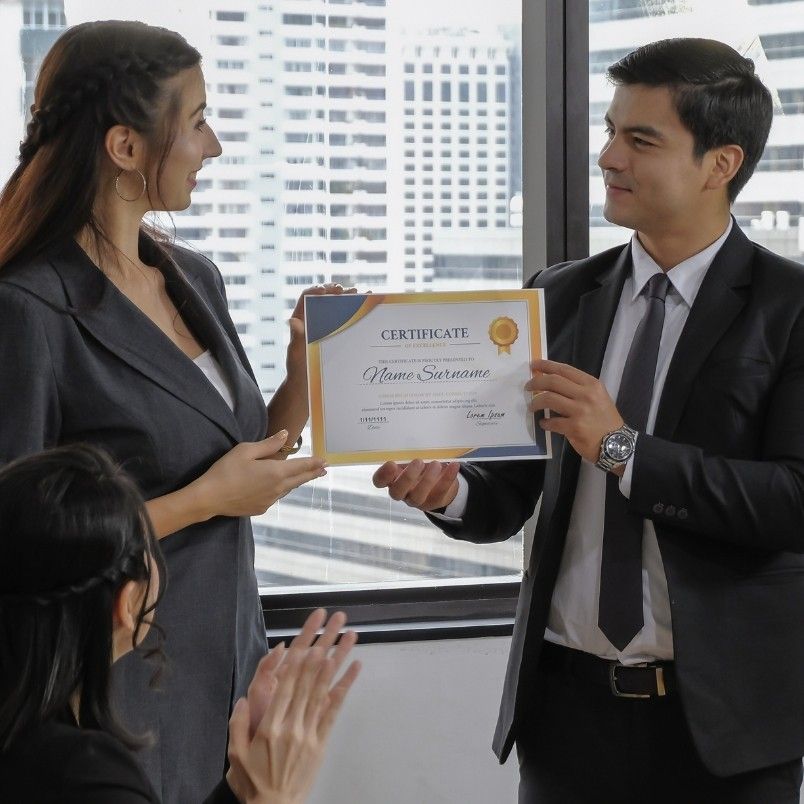A man in a suit hands a certified associate in project management certificate to a woman in professional attire while another person sits nearby clapping. They are indoors with large windows showing city buildings in the background.