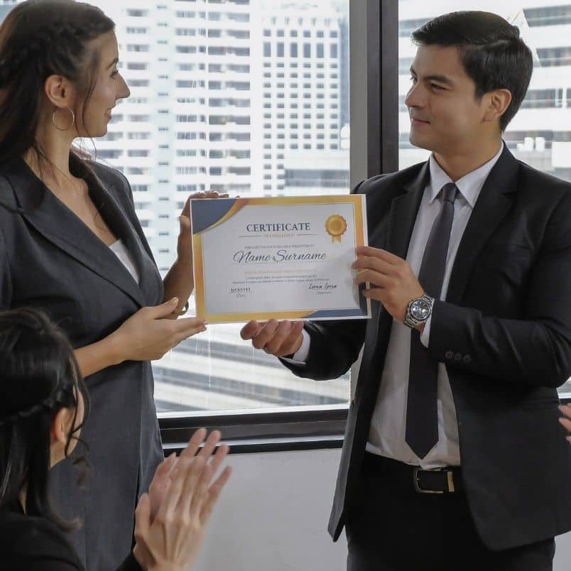 A man in a suit hands a certificate to a smiling woman in business attire, recognized for completing a CAPM course, while colleagues seated nearby applaud. Large windows show a cityscape in the background.