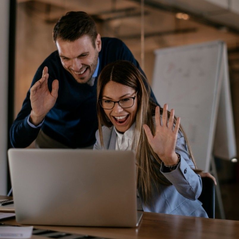 A man and a woman smile and wave at a laptop screen, engaged in agile team collaboration in a hybrid work model, in a modern office setting with a whiteboard in the background.