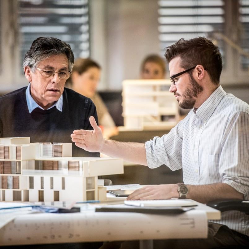 Two men discuss a modern architectural model at a table, with one gesturing toward the model in a collaborative project environment. Papers and notebooks are on the desk, and two blurred people sit in the background.