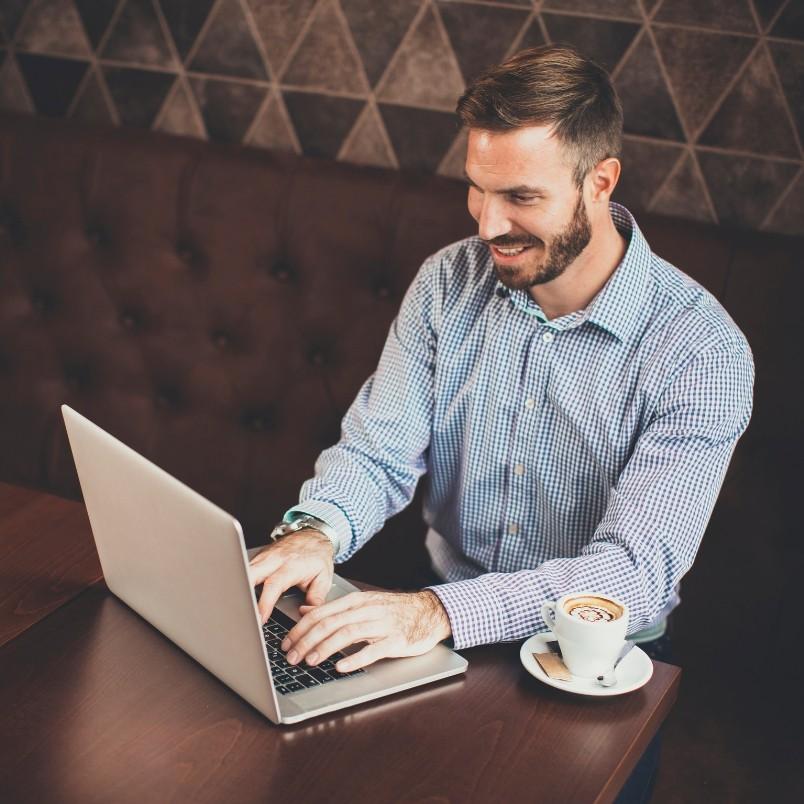 A man with a beard and checked shirt smiles while working on his laptop at a café table, researching generative AI in project management, with a cup of cappuccino beside him.
