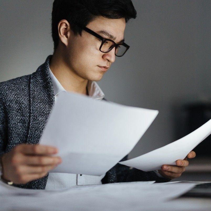 A man wearing glasses and a blazer sits at a desk, focused on reading and sorting through several sheets of paper as part of his PMP exam prep.