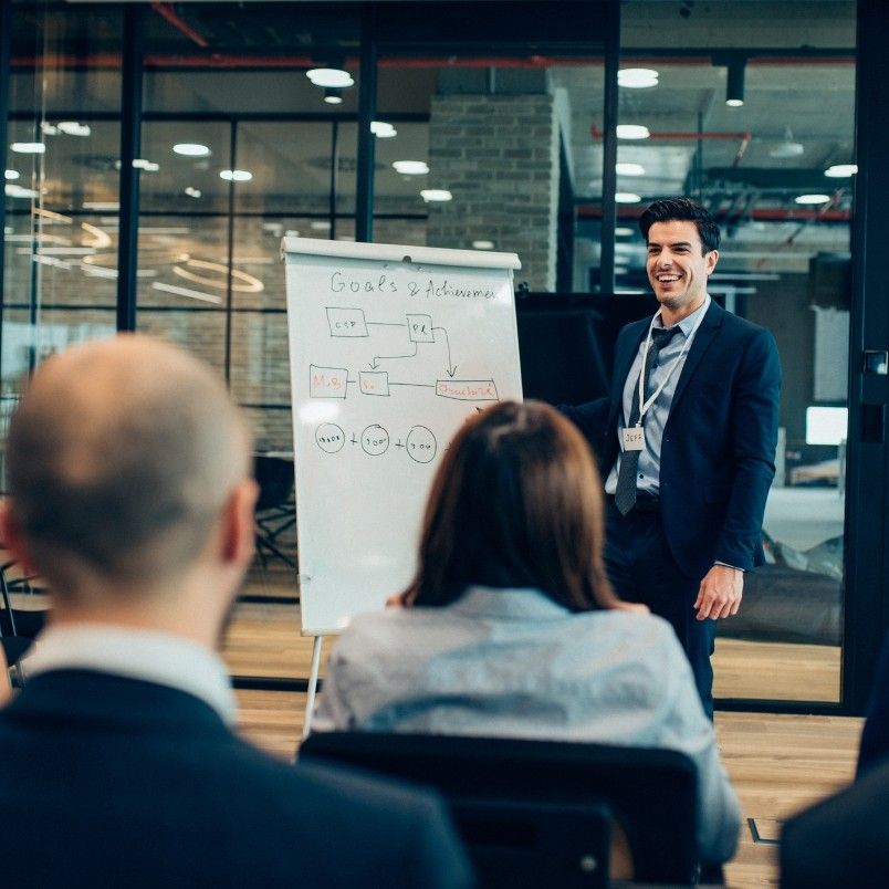 A man in a suit stands by a flip chart, presenting on project governance to an audience in an office setting. The flip chart displays a flowchart labeled "Goals & Achievements," while attendees are seated and listening attentively.