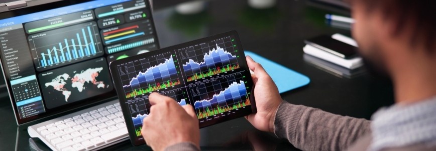 A person analyzes financial data on a tablet displaying four graphs, while a laptop nearby shows charts, graphs, and a world map—suggesting ai-augmented agile metrics and reporting for enhanced financial or stock market analysis.