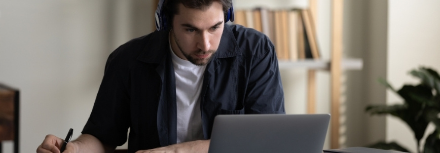 A young man wearing headphones sits at a desk, focused on a laptop screen. He is writing in a notebook, possibly taking notes for a CAPM training course online, with shelves of books and a plant visible in the background.