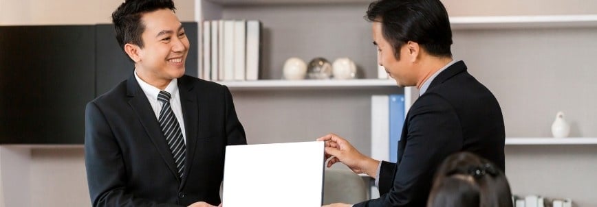 Two men in suits smile while holding a blank white rectangular board together in an office setting, with shelves and decorative items behind them—perfect for presentations on topics like "what is CAPM certification.