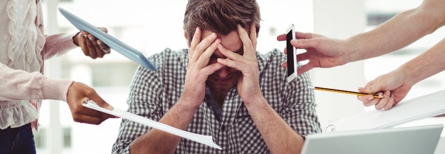 A stressed man sits at a desk with his head in his hands while several people overwhelm him with papers and calls, highlighting the pressure of managing tasks without support from resources like a CAPM training course.