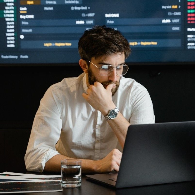 A man with glasses and a beard sits at a desk, looking thoughtfully at a laptop. Papers, a glass of dark beverage, and a large screen displaying ai-augmented agile metrics and reporting are visible in the background.