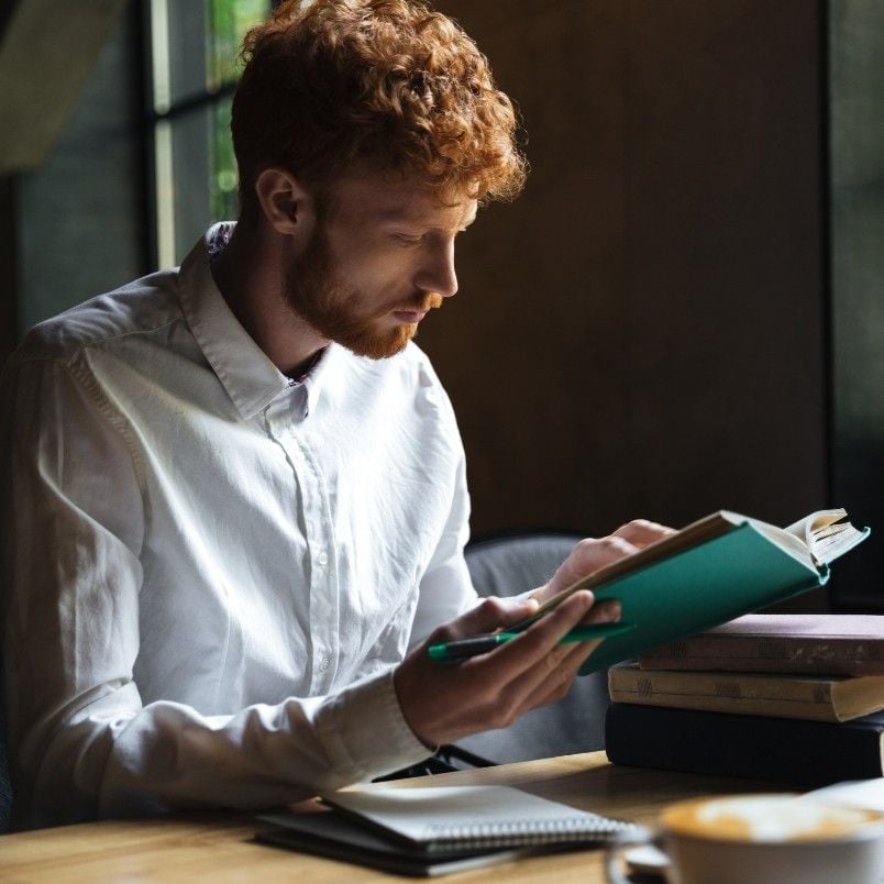 A man with curly red hair and a white shirt sits at a table reading about what is CAPM certification, with a notebook, a stack of books, and a cup on the table in front of him. Natural light filters in from a nearby window.