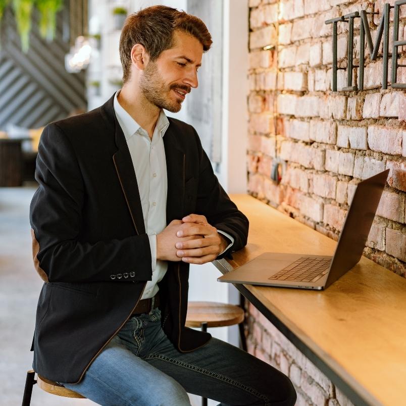 A man in a blazer and jeans sits at a wooden counter, smiling at his open laptop while taking a CAPM training course online. He is indoors, next to a brick wall with decorative lettering in a casual, modern setting.