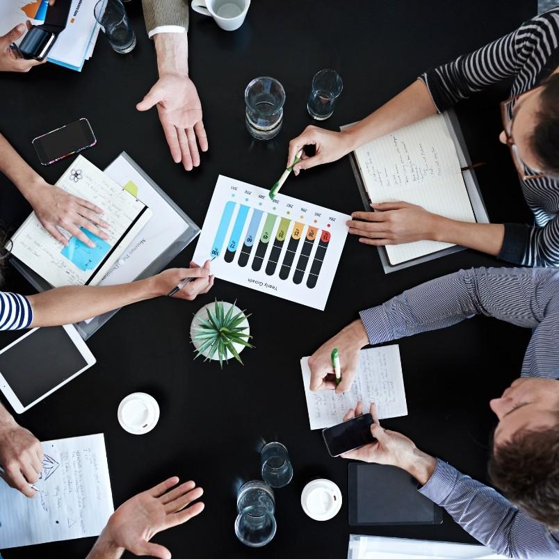 Top-down view of a meeting table with five people’s hands, papers, notebooks, a chart with colored bars reflecting ai-augmented agile metrics and reporting, glasses of water, a small plant, a tablet, and a smartphone—suggesting collaborative work discussion.