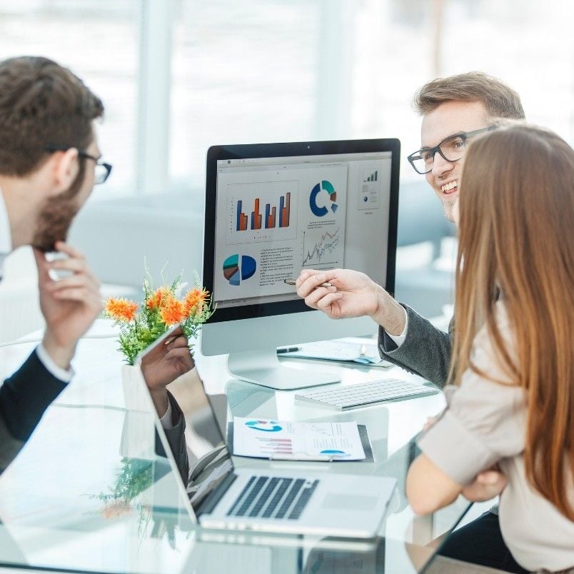 Three people sit around a desk, discussing charts and graphs related to AI-augmented agile metrics and reporting on a computer monitor. Laptops, documents, and a small vase of flowers are on the glass table as they engage in a focused business meeting.