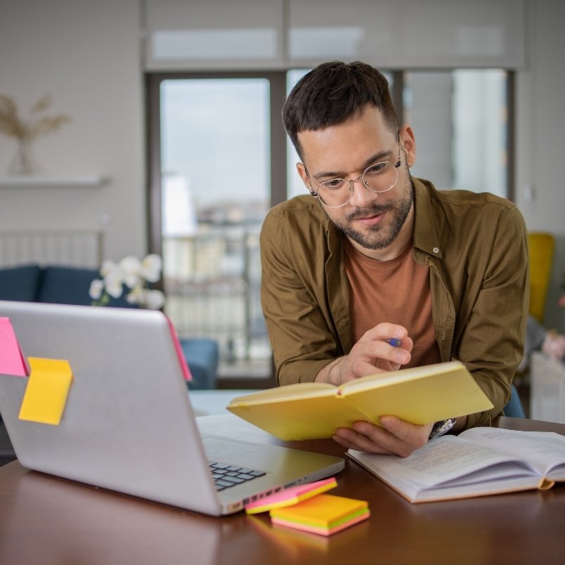 A man with glasses sits at a desk, studying a yellow notebook for his CAPM training course. A laptop covered in colorful sticky notes is in front of him, and more notes are scattered on the desk as he works from home.
