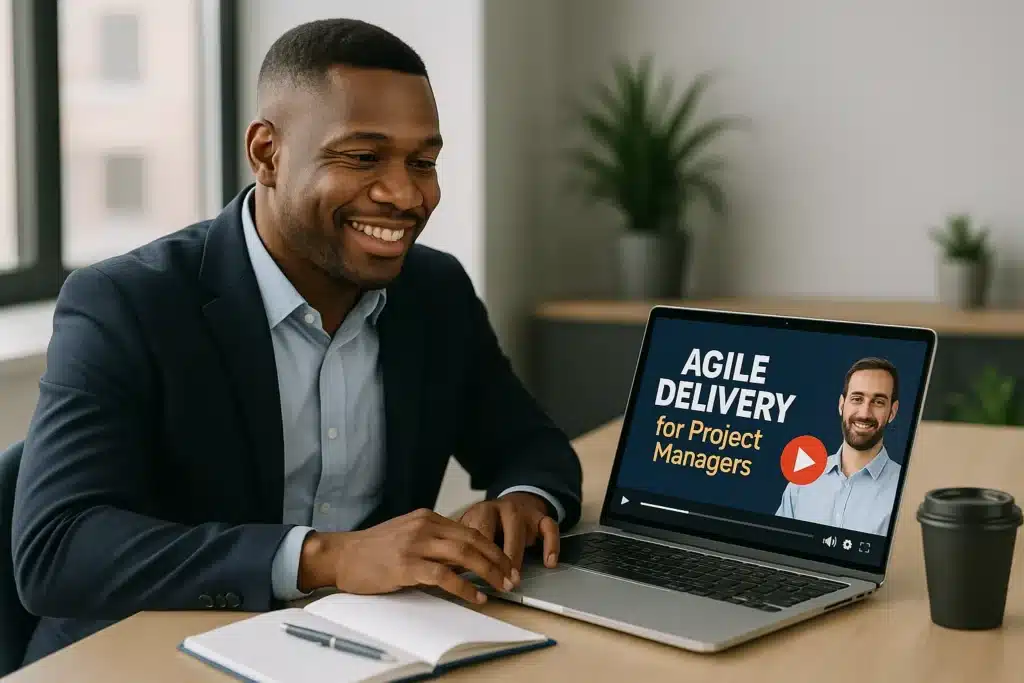 A smiling man in a suit sits at a desk with a notebook, using a laptop displaying an online course titled “AGILE DELIVERY for Project Managers.” A coffee cup and potted plants are in the background.