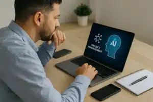 A man sits at a desk using a laptop displaying graphics of a brain labeled "AI" and the words "Project Management." Nearby are a smartphone, a notebook with a pen, and a small potted plant.