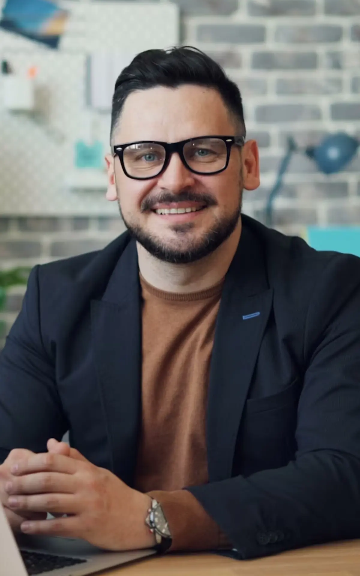 A man with dark hair, a beard, and glasses is smiling while sitting at a desk with a laptop. He wears a brown shirt under a dark blazer. The background features a brick wall and office decor.