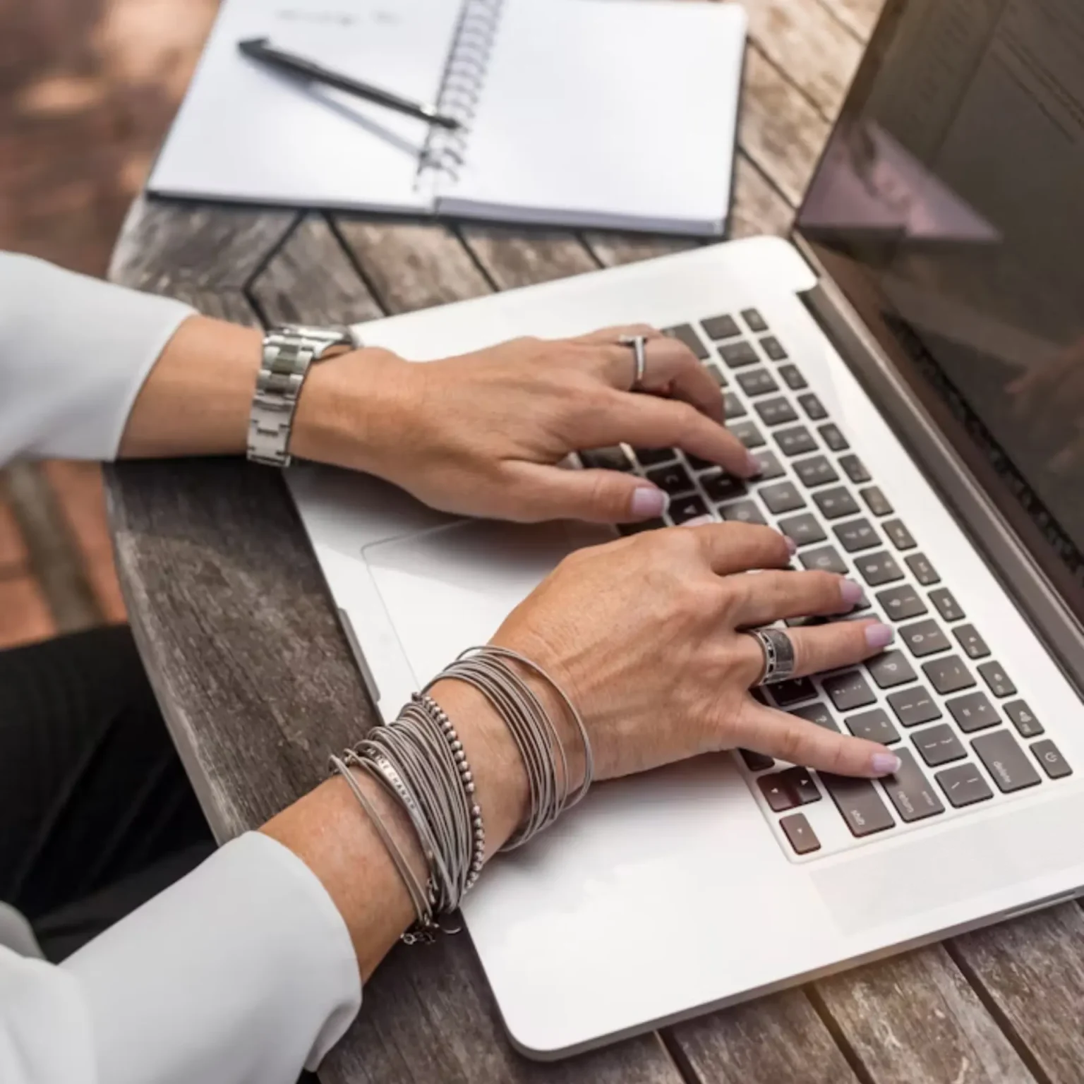 Person with bracelets and rings typing on a laptop at a wooden table, with an open notebook, pen, and smartphone nearby.