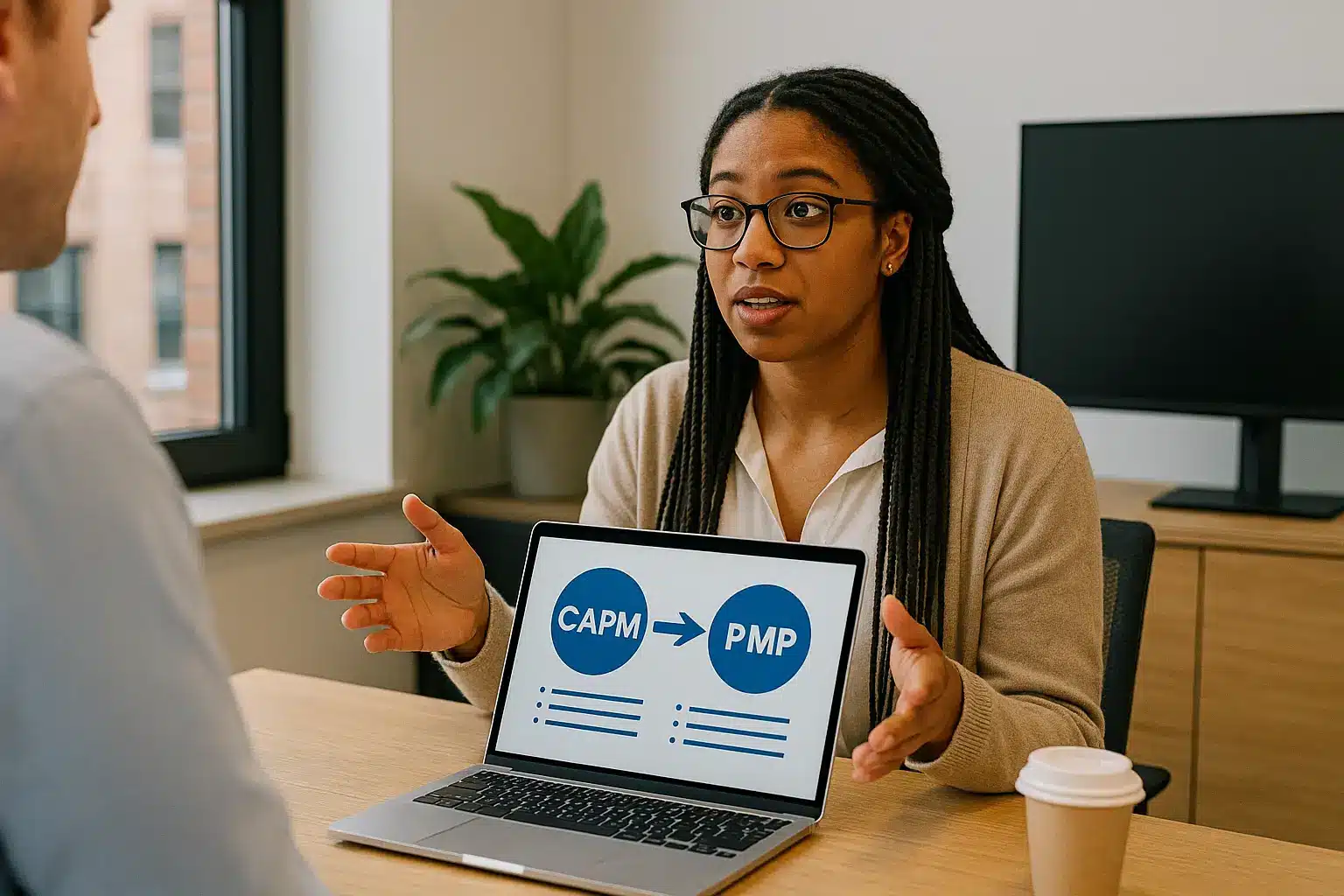 A woman sits at a desk in an office, holding a laptop that displays a diagram showing "CAPM" leading to "PMP." She is talking to a man sitting across from her.