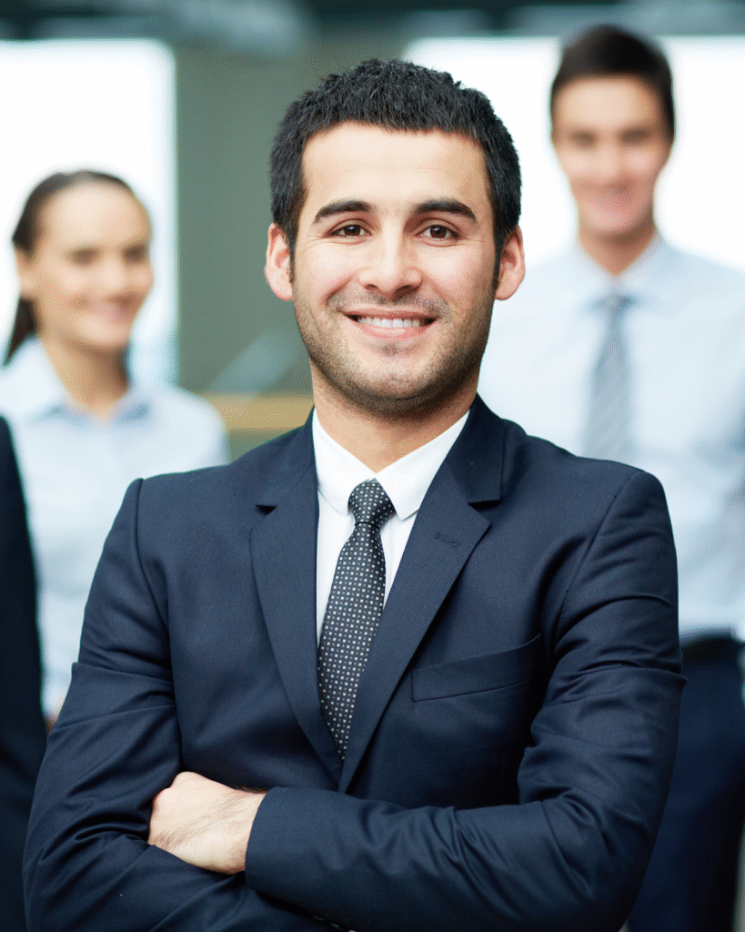 A confident man in a dark suit stands with arms crossed and smiles at the camera, exuding California professionalism, while two blurred colleagues in business attire appear in the background.