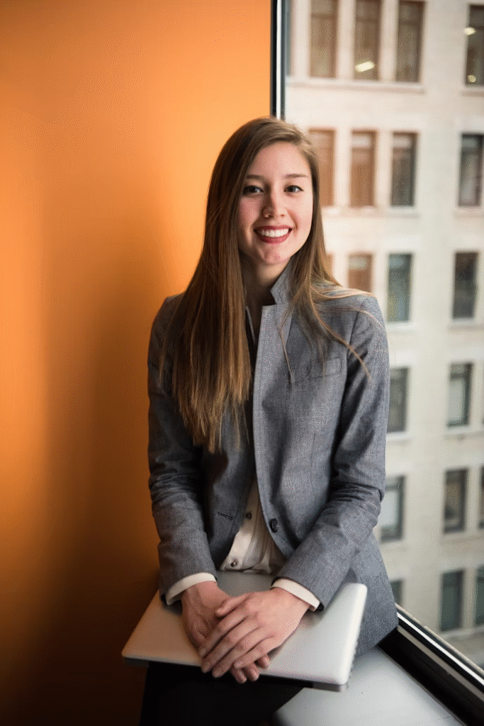 A woman with long brown hair, wearing a gray blazer and white shirt, sits by a window against an orange wall, smiling and holding a closed laptop on her lap—perhaps ready to start her CAPM Course Bundle. An office building is visible outside.