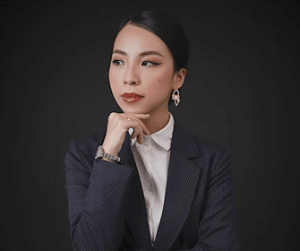 A woman in a dark pinstripe suit and white shirt poses against a black background, resting her chin on her hand and looking thoughtfully to the side. She wears ROSEMET earrings, with styled hair and makeup.