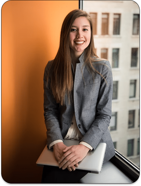 A woman with long brown hair, wearing a gray blazer and white blouse, sits by a window holding a closed laptop on her lap, smiling at the camera. An orange wall is behind her, and a building is visible outside the window.
