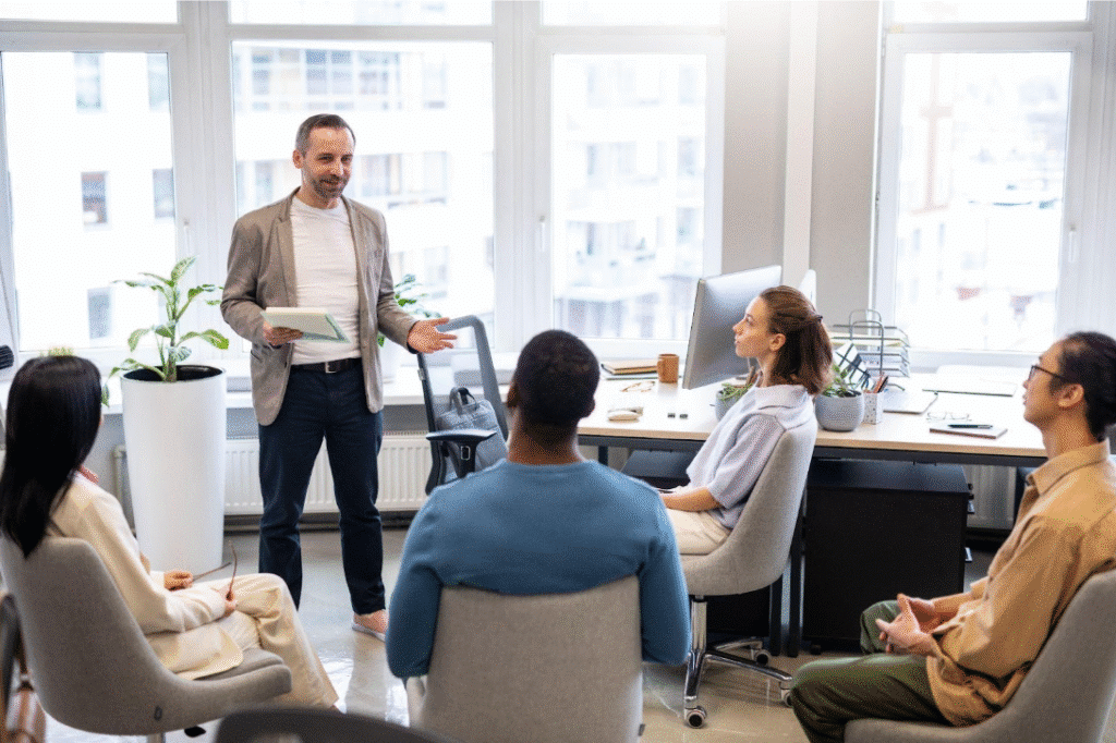 A man stands and speaks to four seated colleagues in a modern office with large windows, desks, and plants. The group appears to be having a discussion or meeting, with everyone focused on the speaker.